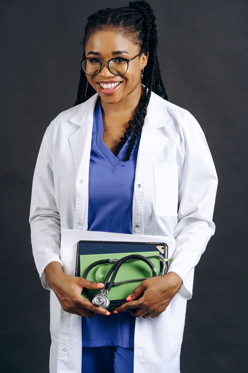 Portrait of a smiling female doctor in a lab coat holding a stethoscope, against a gray background.